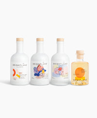 Four Brightland bottles arranged in a row on a clean white background, including three white olive oil bottles (EVOO, lemon flavored, and garlic flavored) with colorful abstract labels and one clear bottle filled with citrus infused champagne vinegar.